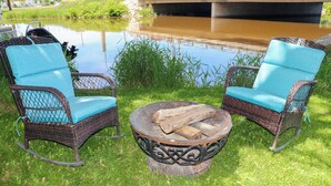 Two wicker rocking chairs with blue cushions, a metal fire pit with logs, and a pond in the background.