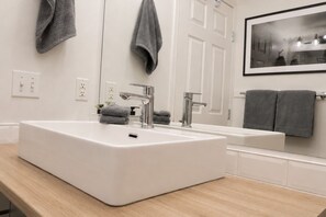 Ein modernes Badezimmer mit einem großen, weißen Waschbecken, zwei Wasserhähnen, einem Spiegel und einem gerahmten Bild an der Wand.