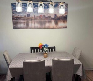 Un coin repas avec une table dressée pour quatre personnes, un tableau représentant un paysage urbain au-dessus, et un vase rempli de fleurs sur la table.