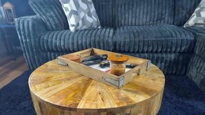 Ein runder Couchtisch aus Holz mit einer Ablage, auf der Fernbedienungen und eine Tasse Kaffee stehen.
