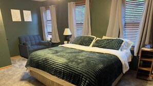 Ein Schlafzimmer mit einem Bett, einem Nachttisch, einer Lampe, einem Sessel und zwei mit Vorhängen versehenen Fenstern.