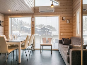 Interior de una cabaña de madera con comedor, sofá y ventanas amplias que ofrecen vistas de otra cabaña y árboles.