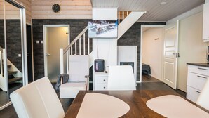 Uma sala de estar moderna com uma mesa de jantar de madeira, cadeiras brancas, televisão fixada na parede, uma escada e uma área de cozinha.