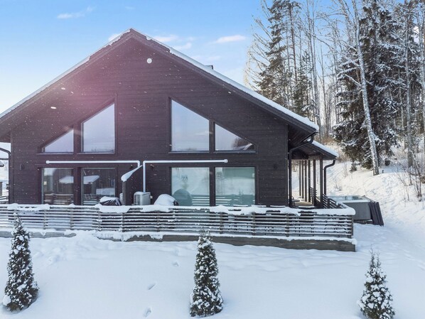Una cabaña moderna de madera con ventanas amplias, un porche cubierto y un paisaje cubierto de nieve.