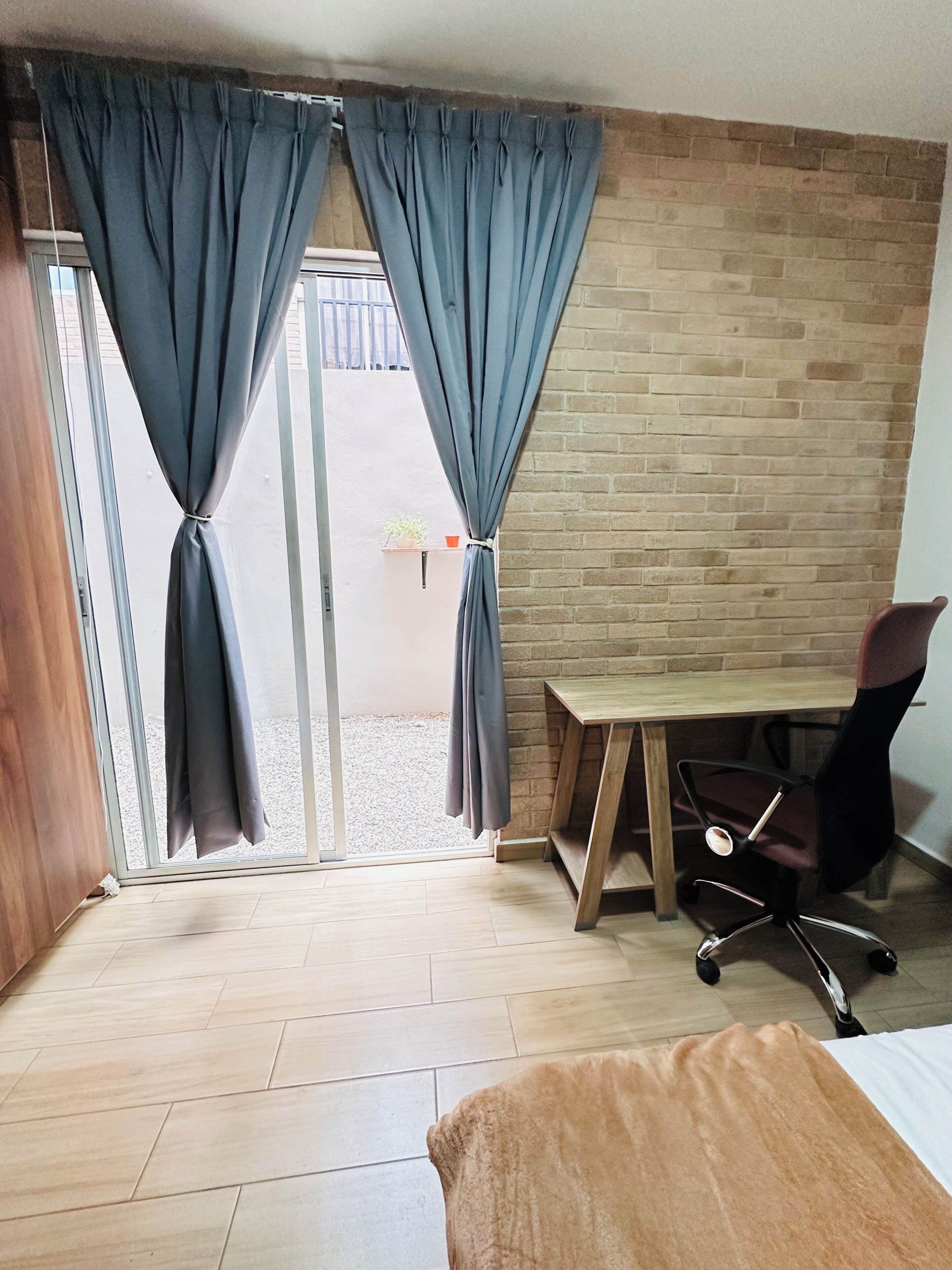 Une pièce avec un bureau en bois, une chaise, un lit et une vue sur l’extérieur, visible à travers une porte vitrée munie de rideaux.