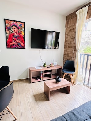 Ein modernes Wohnzimmer mit einem Flachbildfernseher, einem Holztisch, einer schwarzen Couch, einem schwarzen Sessel und einem farbenfrohen Bild an der Wand.