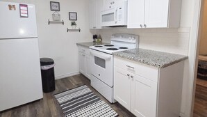 A compact kitchen with white cabinetry, a microwave, a stove, and a refrigerator. There are framed pictures on the wall and a small trash bin.