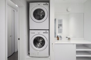 Un área de lavandería compacta con dos lavadoras apiladas, un lavabo blanco y un espejo sobre la encimera.