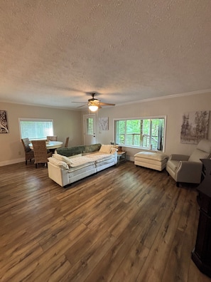 A spacious living room with hardwood flooring, a sectional sofa, a dining area with a table and chairs, and a ceiling fan.