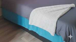 Ein Schlafzimmer mit einer gepolsterten Kopfteile, einem Bett mit blau-weißer Bettwäsche, einer grauen Decke und einem Fenster mit einem weißen Vorhang.
