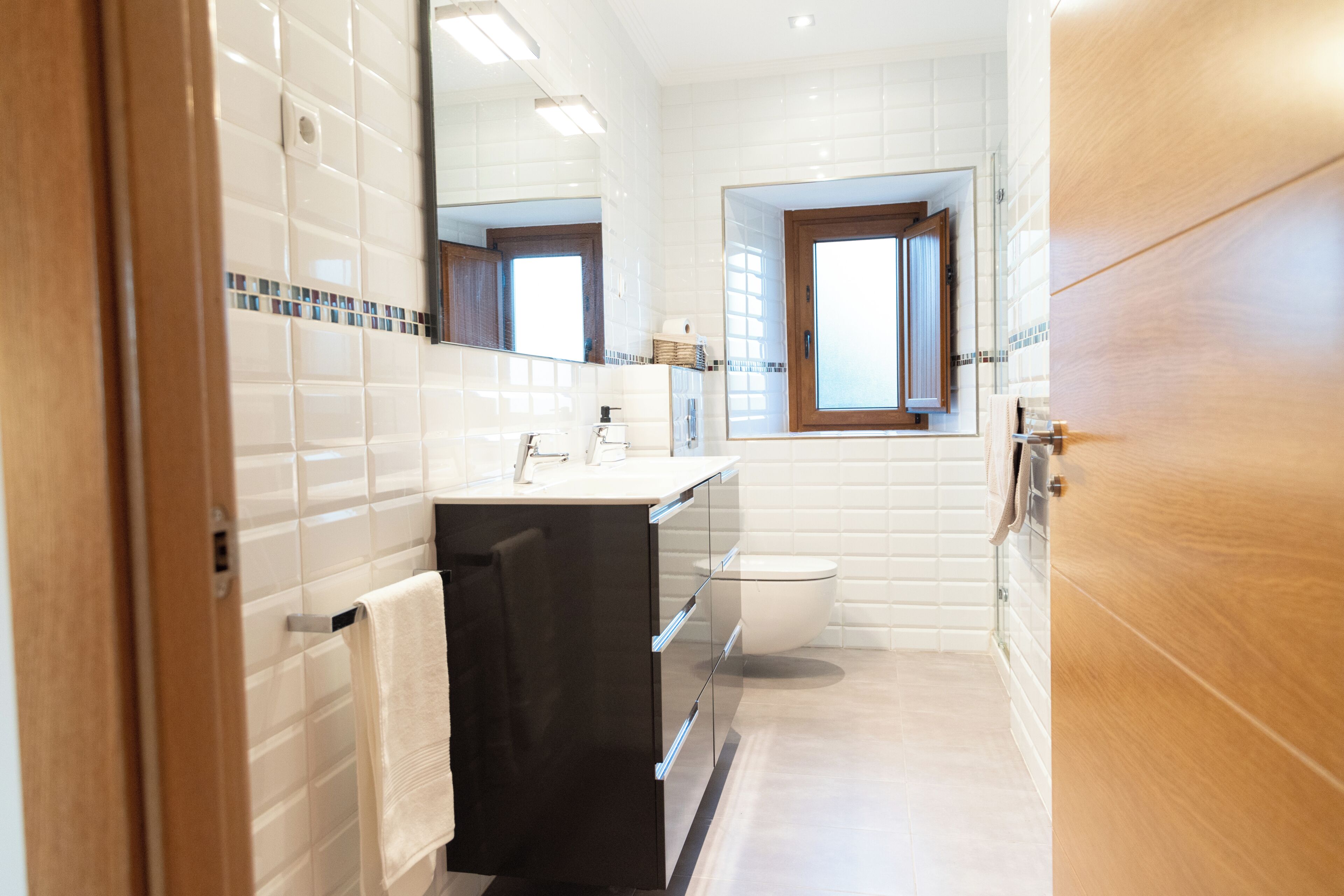 Un baño moderno con azulejos blancos, un tocador negro, cabina de ducha de vidrio y puerta de madera.
