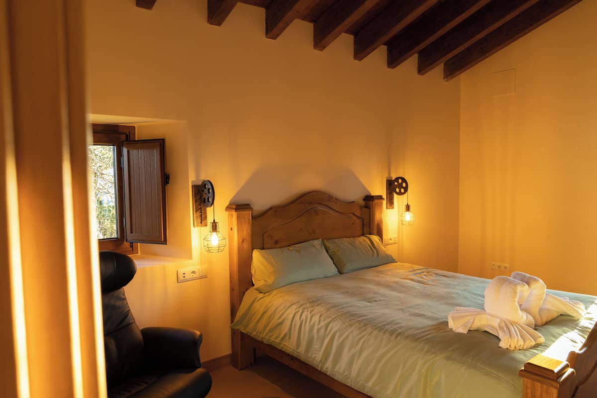 A bedroom with a wooden headboard, a bed with a green bedspread, two wall-mounted light fixtures, and a window with curtains.