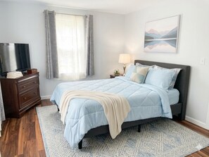 Quarto com cama, penteadeira, luminária, televisão e janela com cortinas.