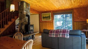 A cozy cabin interior with a stone fireplace, wooden ceiling, and a large window overlooking a forest. - Off Grid Retreat Stay for 7 (Rexford)