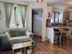 A compact living space with a sofa, a small table, a kitchen area with a sink and stools, and a doorway leading to another room.