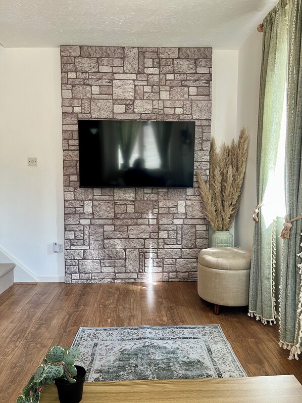 A living room with a stone wall, a flat-screen TV, a wooden floor, a patterned rug, and a potted plant.