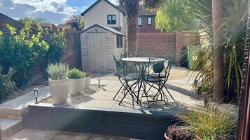 Un espace patio aménagé avec une table et des chaises, entouré de plantes en pot et d’un petit abri de jardin.