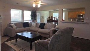 A living room with a sectional sofa, a coffee table, and a chair. There is a ceiling fan, a dining area with chairs, and a kitchen area with cabinets and appliances. - Hook, Line & Relax – Coastal TX Getaway (Texas City)
