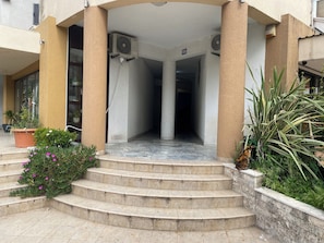 Entrada de un edificio con escalones que conducen a un área techada, unidades de aire acondicionado y plantas en macetas.
