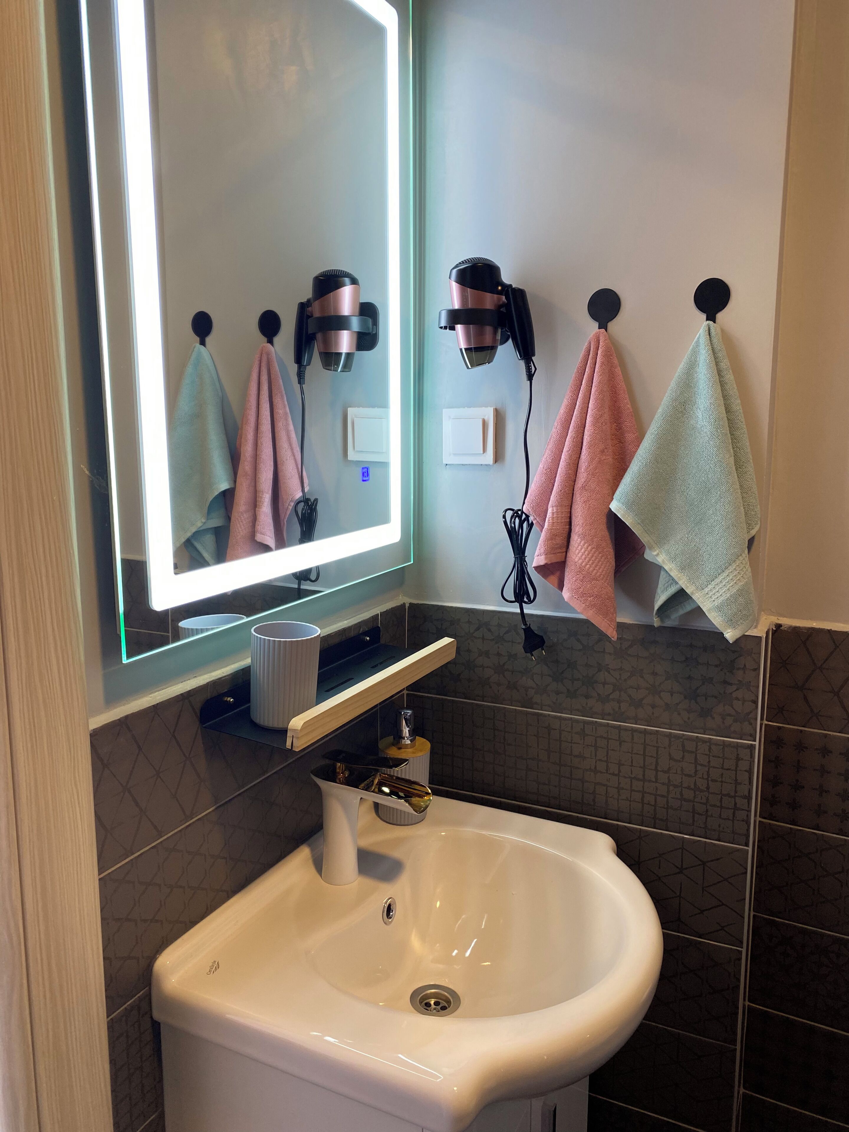A bathroom with a white sink, a mirror with lights, and towels hanging on hooks.