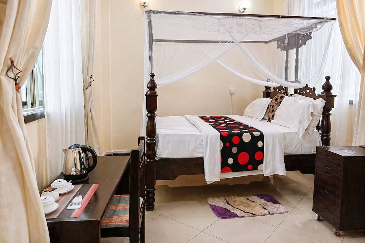 Una cama con dosel, una mesita de noche con hervidor, un armario pequeño y una alfombra estampada.