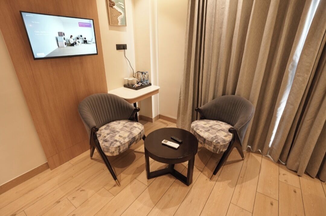 Quarto de hotel com duas poltronas, uma mesa pequena, televisão na parede e cortina.