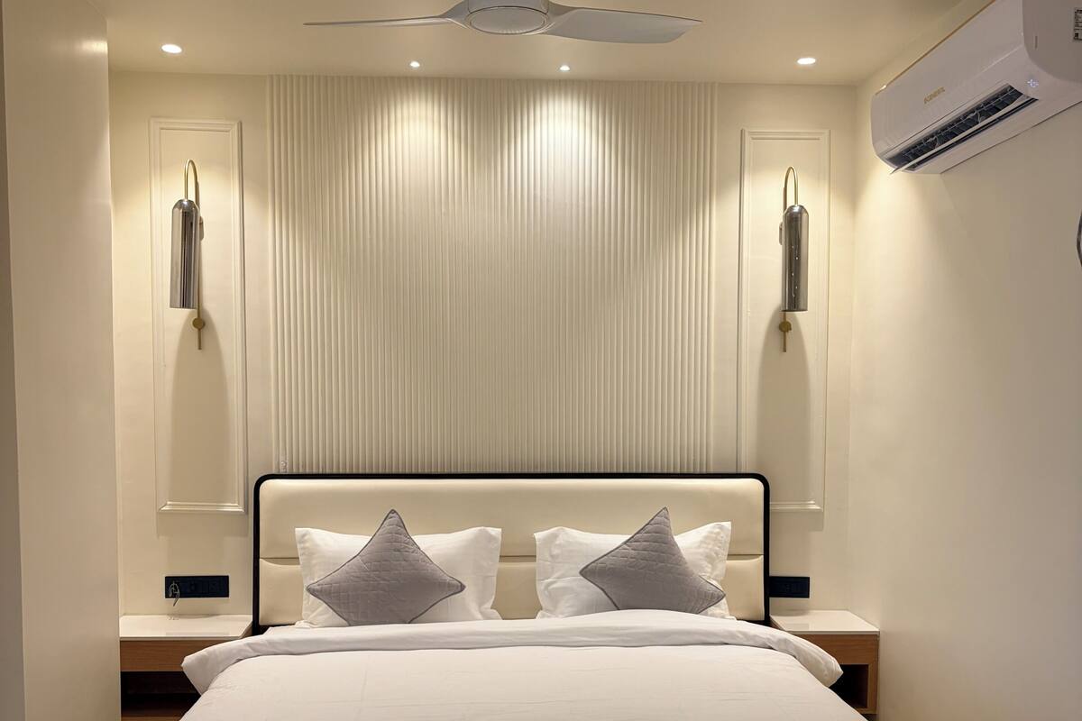 A modern hotel room with a large bed, a ceiling fan, an air conditioning unit, and a wall-mounted light fixture.
