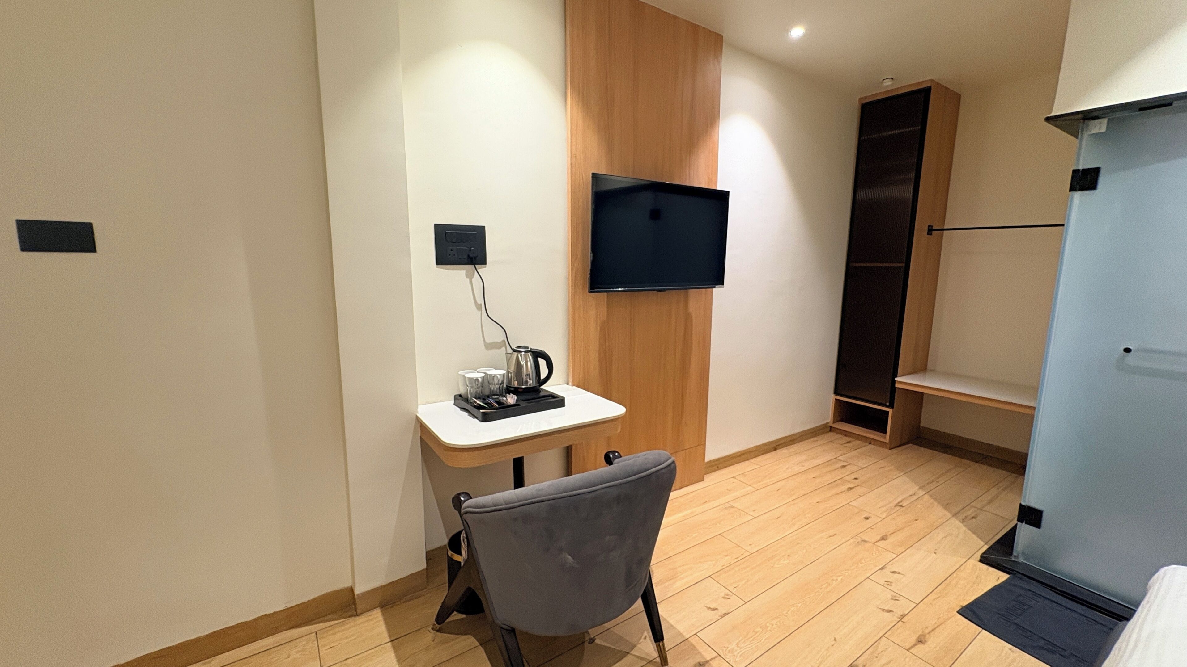 A small room with a wooden panel wall, a flat-screen TV mounted above a desk, a chair, and a wardrobe with a mirror.