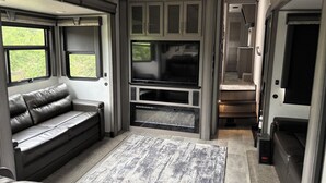 Ein kompakter Wohnraum mit Sofa, Flachbildfernseher, Kamin und Fenster mit Blick auf Grünflächen.