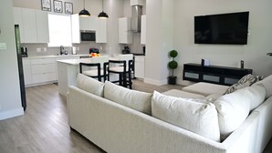 Eine moderne Küche mit weißen Schränken, ein Essbereich mit einem weißen Tisch und schwarzen Stühlen, ein beiges Sofa, ein Flachbildfernseher und ein kleiner Topfpflanze.