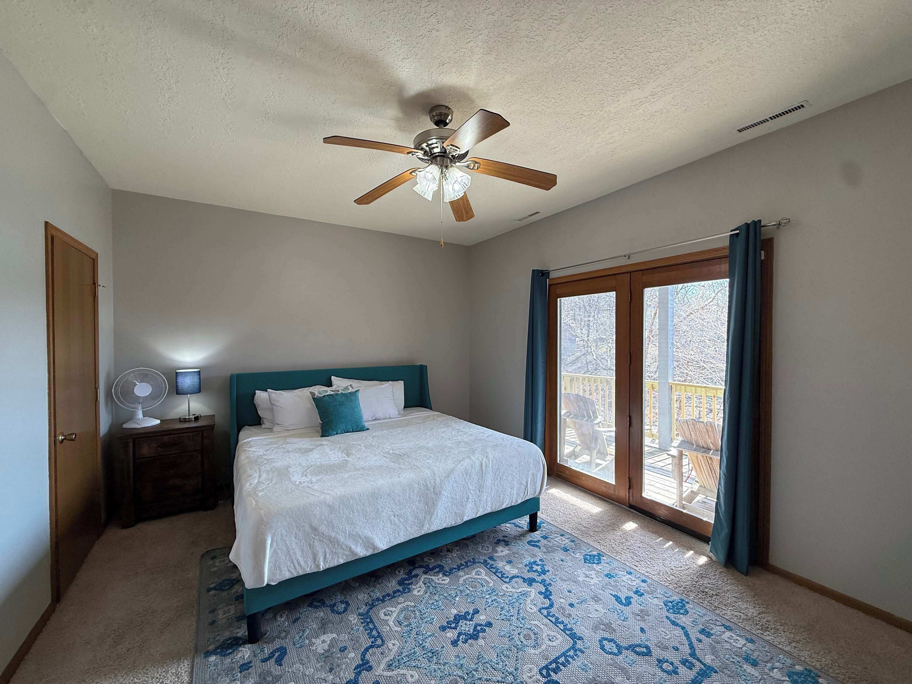 A bedroom with a bed, a ceiling fan, a nightstand, a lamp, a patterned rug, and a sliding glass door to a balcony.