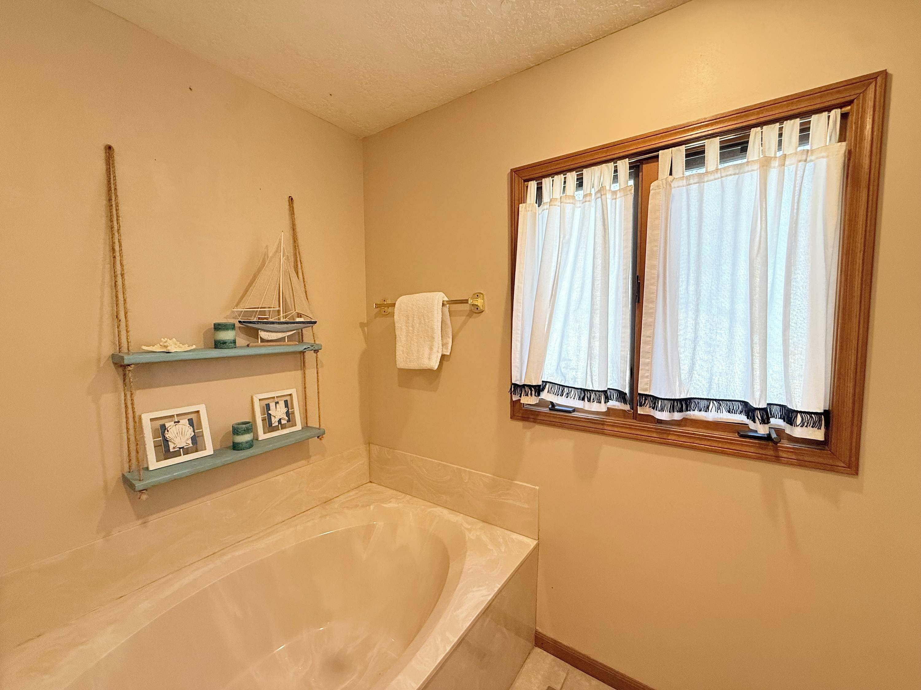 A bathroom with a bathtub, a window with curtains, and a shelf with decorative items.