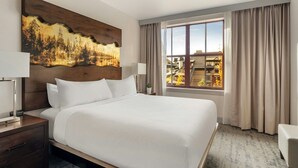 A hotel room with a large bed, a nightstand with a lamp, a painting on the wall, a window with curtains, and a view of buildings outside. - Beautiful spacious 2-bedroom condo in fabulous South Lake Tahoe (South Lake Tahoe)