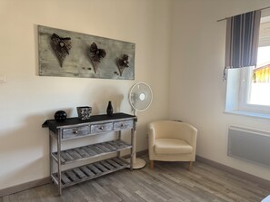 Ein Zimmer mit Holzboden, einem weißen Stuhl, einem Konsolentisch mit Schubladen, einem Wandventilator und einem Fenster mit Vorhängen.
