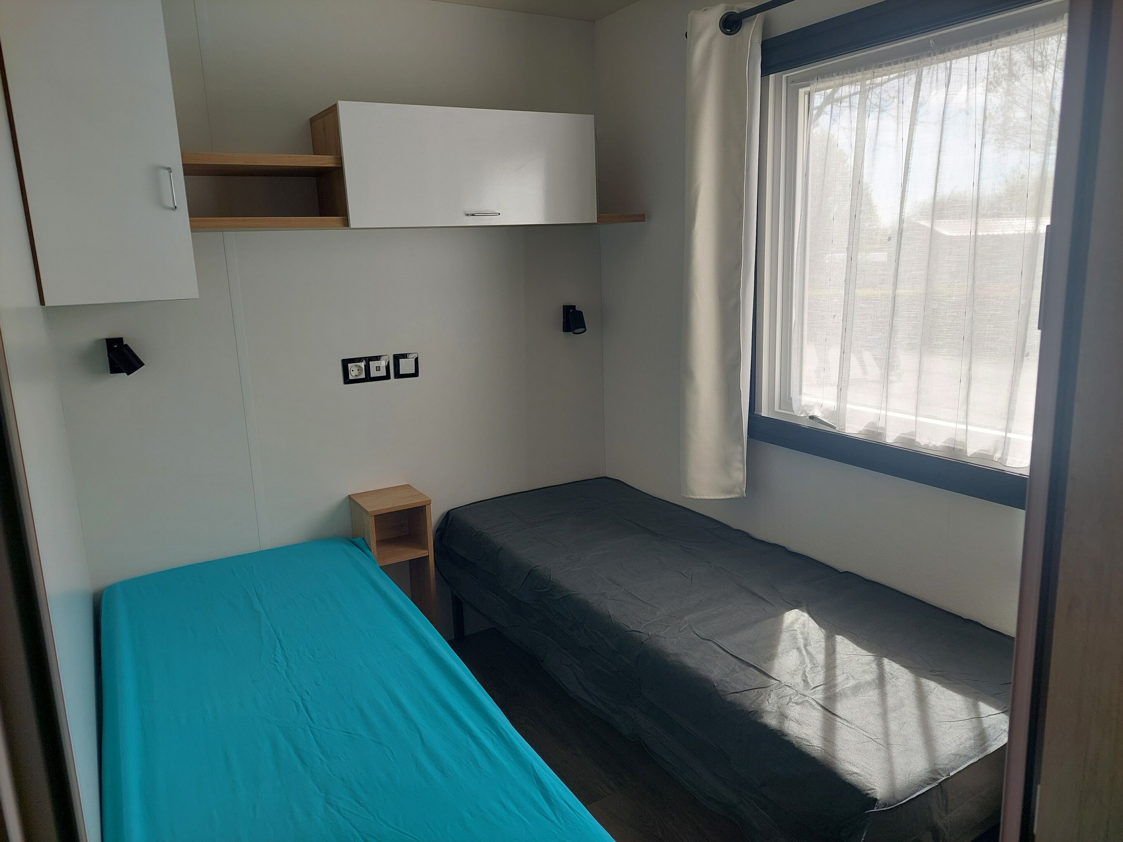 Une petite chambre avec un lit simple, une fenêtre, une table de chevet, une armoire et des prises électriques au mur.