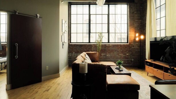 Een moderne woonkamer met een bank, een poef en een televisie. Er is een groot raam met uitzicht, een plant en een bakstenen muur.