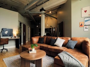 A modern living room with a brown leather sofa, a round wooden coffee table, and a desk with a chair. - Cooperage 214 | 2BR Industrial Downtown Loft (Peoria)