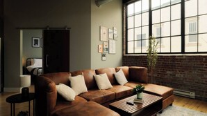 Een moderne woonkamer met een bruine leren bank, een houten salontafel en een groot raam met uitzicht op een bakstenen gebouw.
