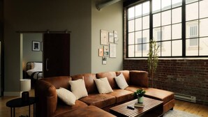 A modern living room with a brown leather sofa, a coffee table, and a large window with a view of a brick building. - Cooperage 214 | 2BD Boutique Loft Stay (Peoria)