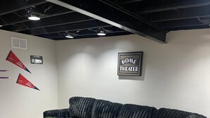 A black sectional sofa with a wooden floor and a sign that reads 'HOME THEATER' on the wall.