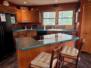 A kitchen with wooden cabinets, a central island, bar stools, a refrigerator, and a window with a view of trees.