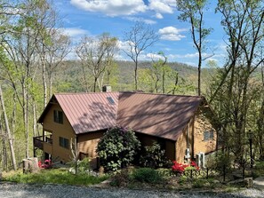 A two-story wooden cabin with a sloped roof, surrounded by trees and a gravel driveway.