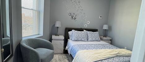 Ein Schlafzimmer mit Bett, Stuhl, Wandbild und einem Fenster mit Jalousien.