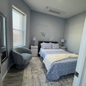 Une chambre à coucher comprenant un lit, une chaise, une œuvre d’art murale et une fenêtre avec des stores.