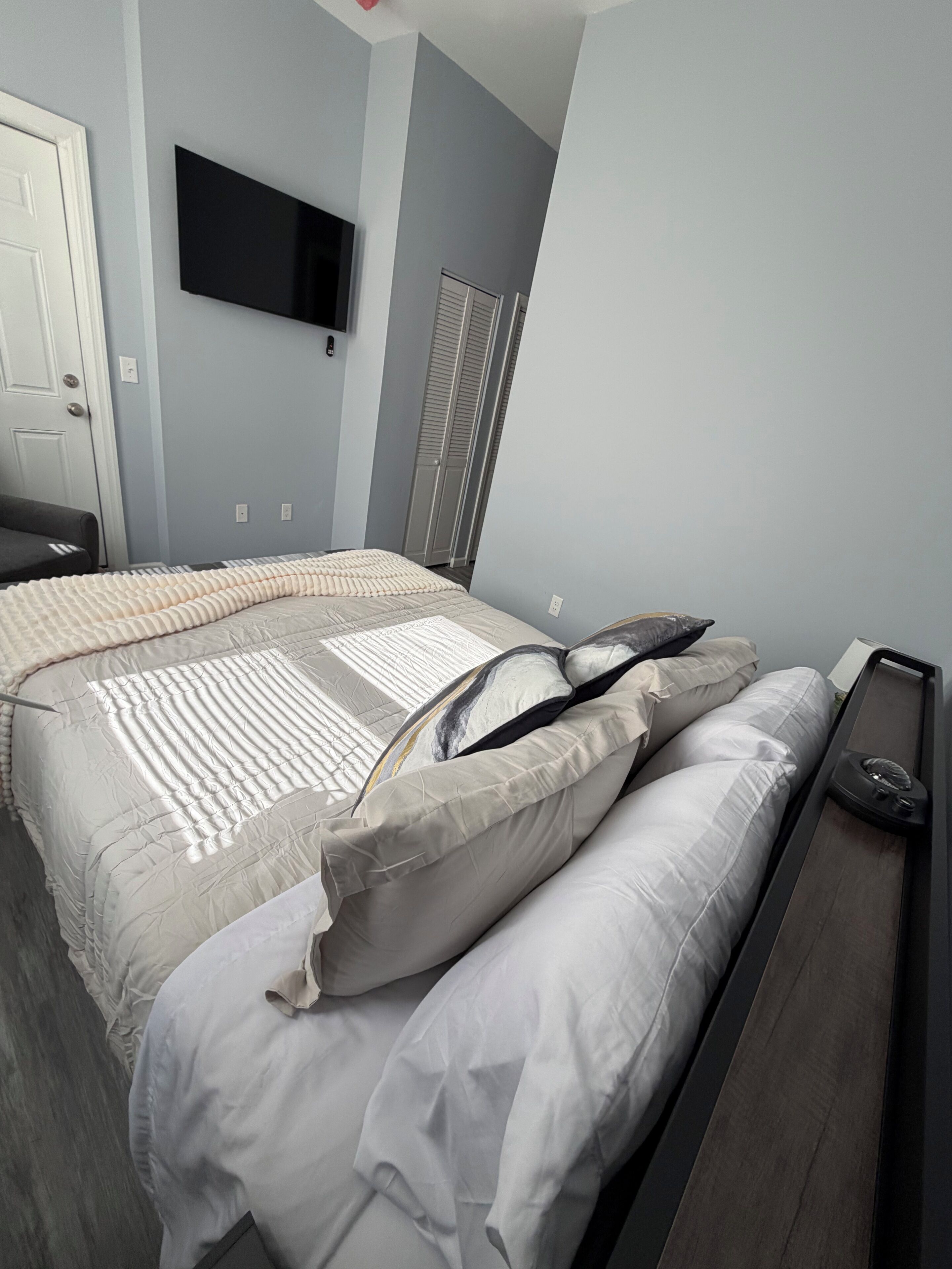 Un dormitorio con cama, mesita de noche, televisor montado en la pared y una puerta.