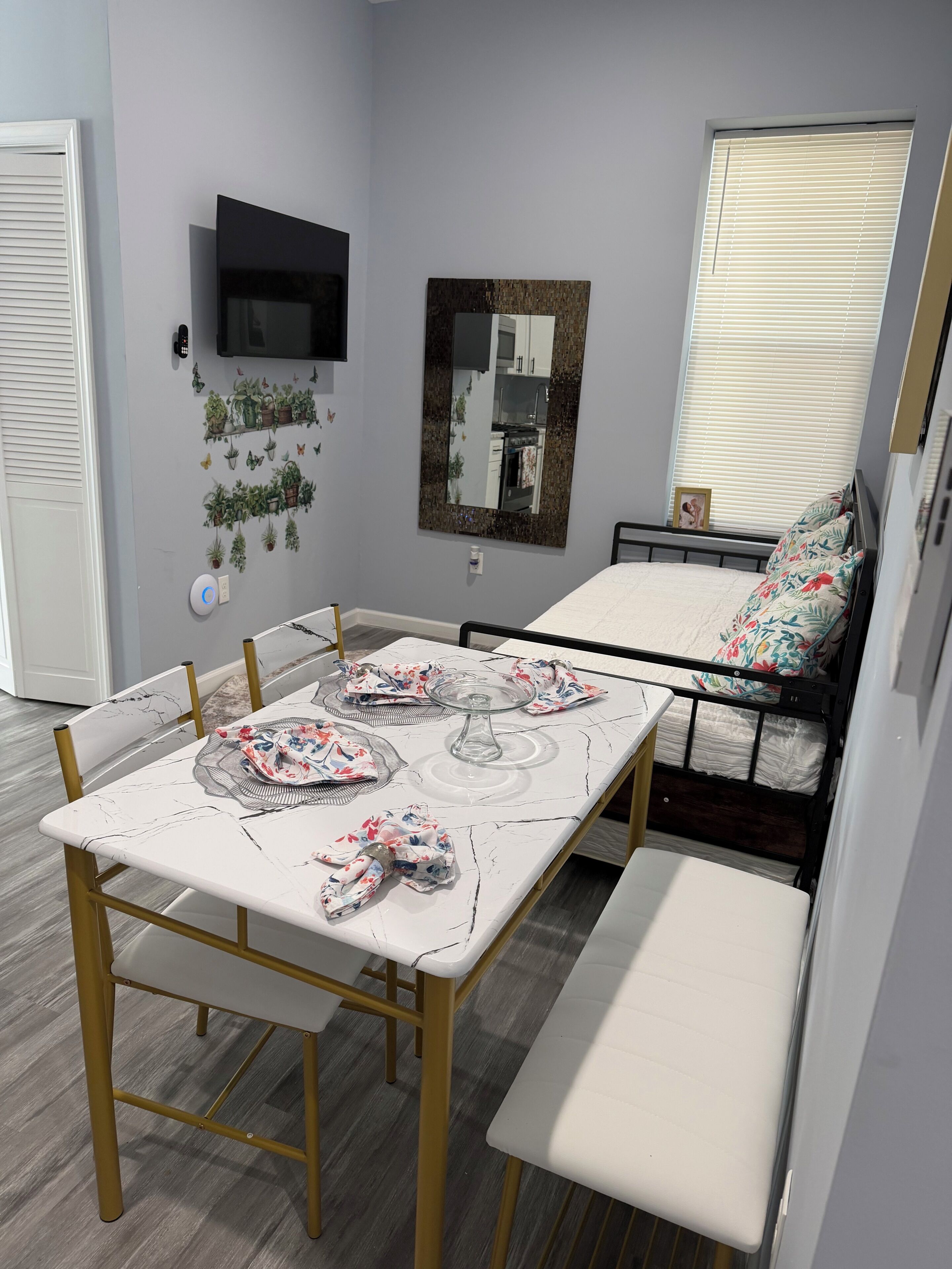 Una habitación compacta con una mesa de comedor, banco, cama, televisor, espejo y plantas colgantes.
