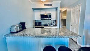 A kitchen with granite countertops, white cabinets, a microwave, and a toaster. There are two black barstools in front of the counter. - Sandy Seawall Escape Resort (Galveston)