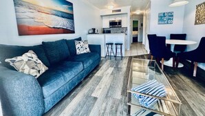 A living room with a sofa, a glass coffee table, a painting, a kitchen area, and a dining table with chairs. - Sandy Seawall Escape Resort (Galveston)