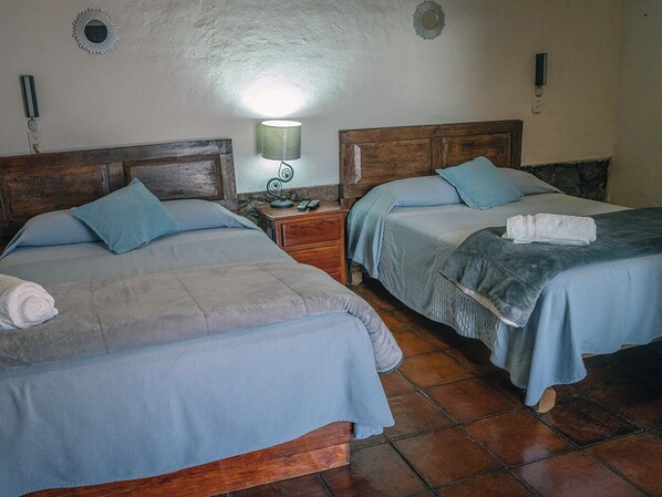 Una habitación con dos camas, cada una con cabecera de madera y colcha azul. Hay una mesita de noche con una lámpara entre las camas.