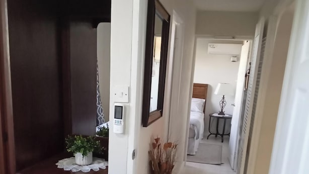 Un pasillo con un armario de madera, una planta en maceta, un candado digital en la pared, un espejo y una cama al fondo.